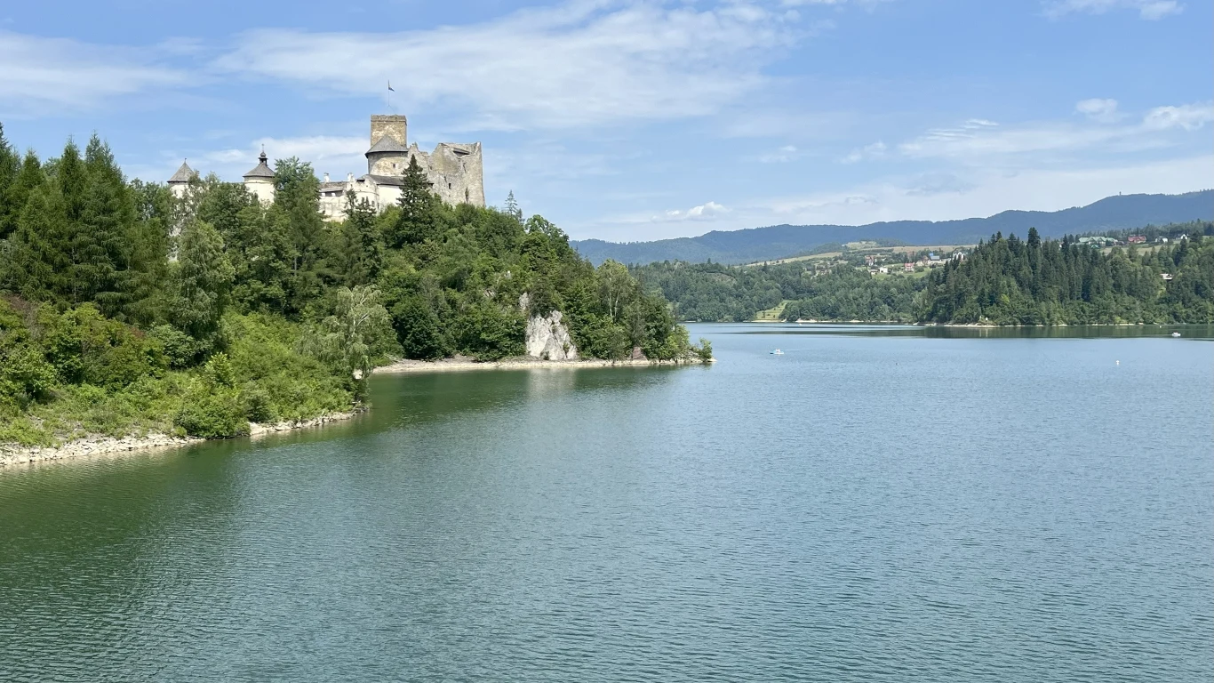 Zamek na wzniesieniu otoczony lasami nad malowniczym jeziorem, w tle góry i rozległe zielone tereny