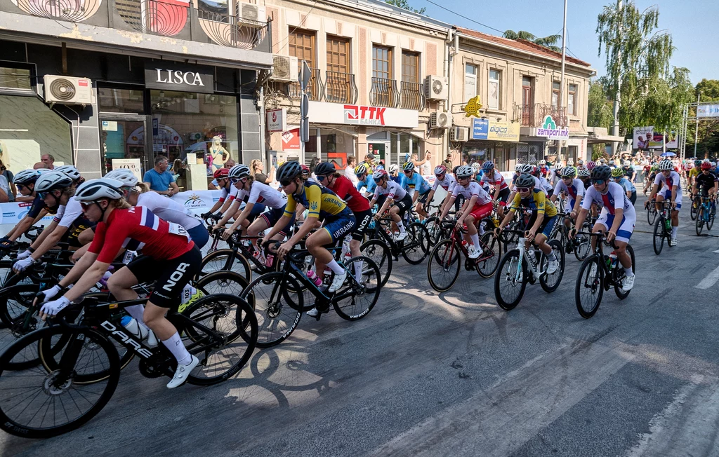 Grupa zawodowych kolarzy bierze udział w wyścigu ulicznym, ścigając się po miejskiej drodze na tle starych budynków handlowych, część rowerzystów ubrana w kolorowe stroje sportowe.
