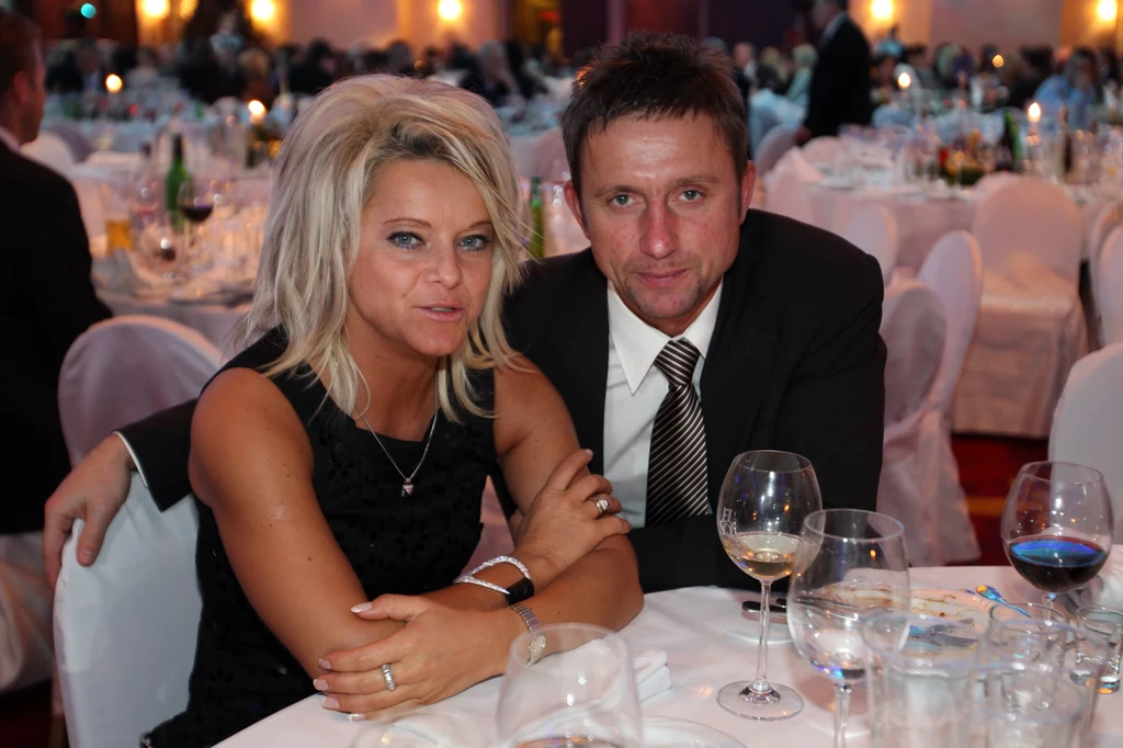 Marzena i Jerzy Brzęczkowie Para elegancko ubranych osób siedzi przy stole na bankiecie, wokół nich widać innych gości oraz zastawione stoły z kieliszkami i talerzami.