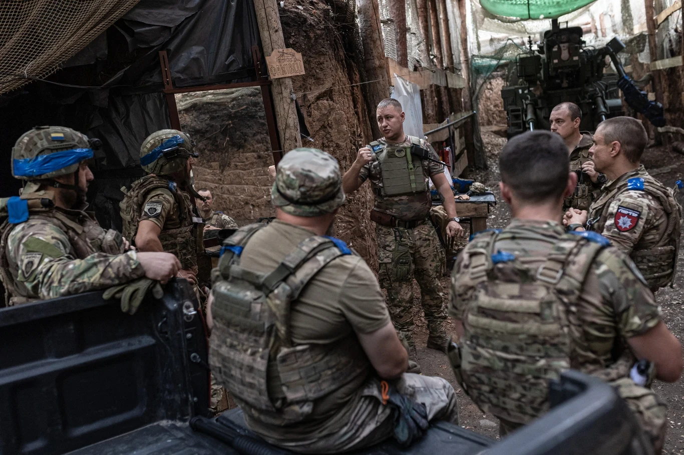 Starlink pomaga Ukraińcom utrzymać łączność na froncie Grupa żołnierzy w pełnym umundurowaniu zgromadzona w okopie, jeden z nich przemawia do pozostałych, wszyscy wyposażeni w kamizelki taktyczne i ochraniacze, wokół widać ziemne ściany i sprzęt wojskowy.