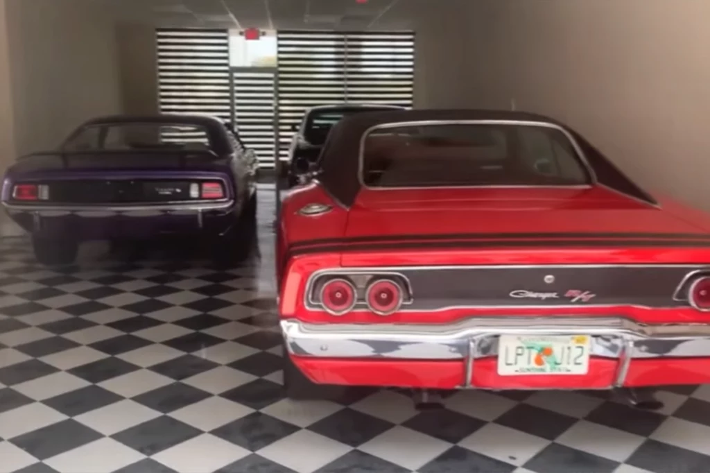 Amerykańskie muslce cary'y w garażu Hulka Hogana. Dwa klasyczne amerykańskie samochody, czerwony Dodge Charger R/T oraz ciemnofioletowy samochód muscle car, zaparkowane na czarno-białej szachownicy podłogowej w zamkniętym pomieszczeniu z przeszklonym wejściem.