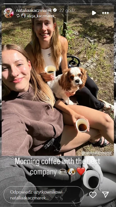 Dwie uśmiechnięte młode kobiety siedzą na zewnątrz, trzymając filiżanki z kawą, między nimi znajduje się mały pies rasy cavalier king charles spaniel, w tle widać fragment ogrodu i promienie słońca.