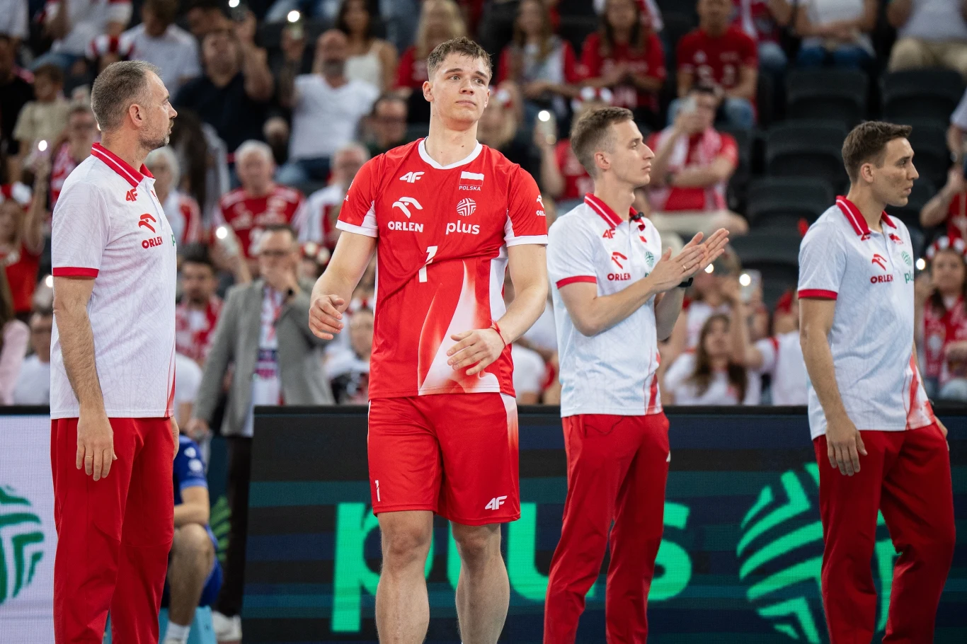 Zawodnik siatkarski w czerwonym stroju stoi na boisku w towarzystwie trzech członków sztabu szkoleniowego ubranych w biało-czerwone koszulki, w tle widownia kibicująca drużynie.