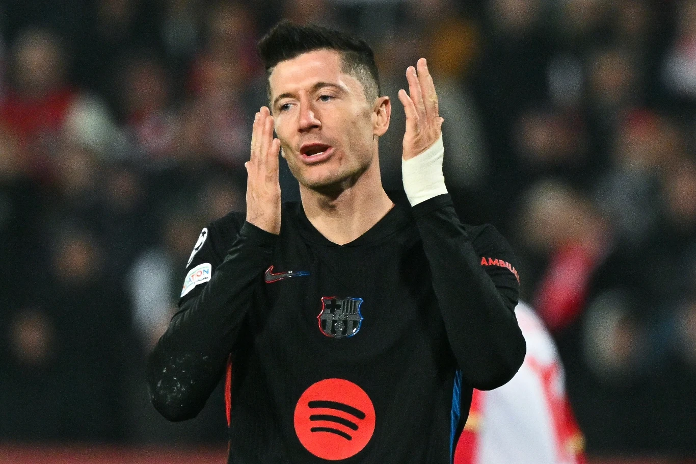 Robert Lewandowski Piłkarz w czarnym stroju FC Barcelony z wyraźnym logiem sponsora, z bandażem na lewej dłoni, wykonuje gest rękoma w stronę twarzy, w tle rozmyte sylwetki oraz trybuny stadionu.
