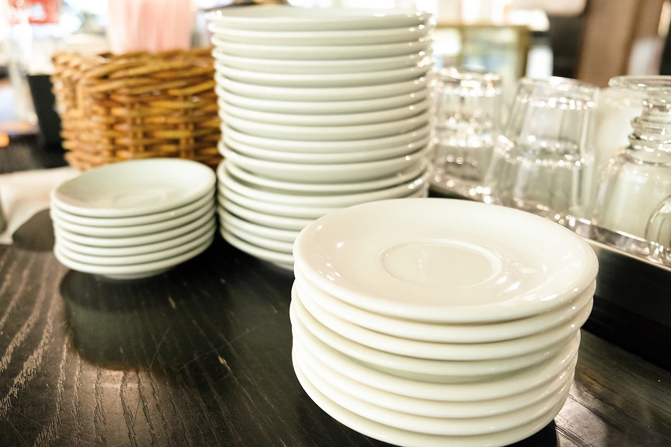 GIS wydał ostrzeżenie publiczne dotyczące wyrobu do kontaktu z żywnością / zdjęcie ilustracyjne Stosy białych, porcelanowych spodeczków ustawione na ciemnym blacie, w tle wiklinowy koszyk oraz odwrócone szklanki.