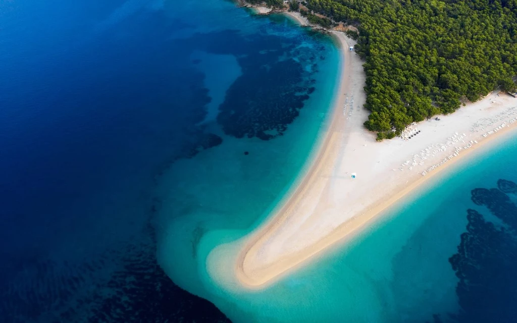 Wąski, jasny pas piaszczystej plaży otoczony turkusową wodą i bujnym, zielonym lasem, widziany z lotu ptaka, miejsce przejścia plaży w morze tworzy charakterystyczny cypel.
