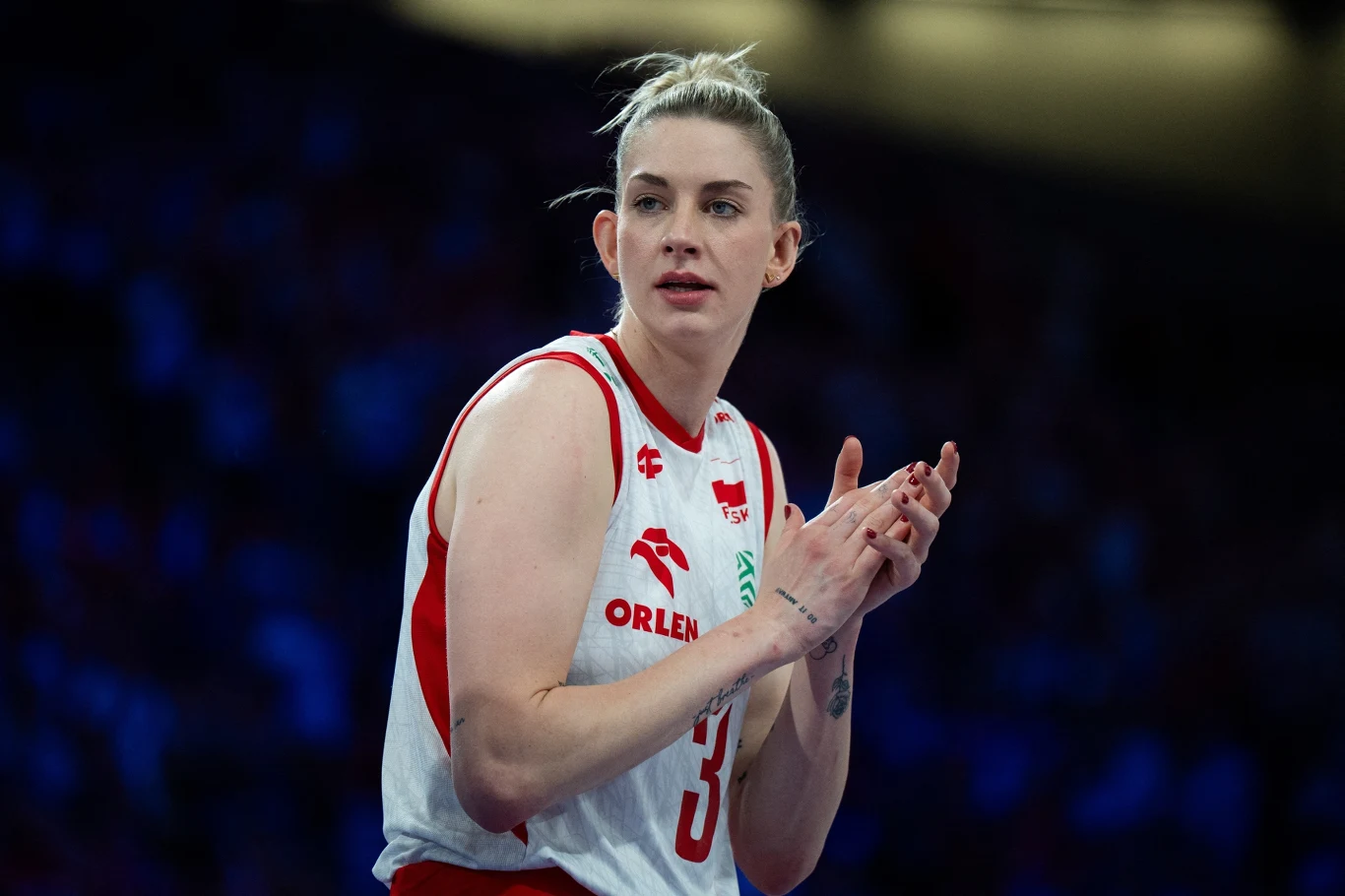 Koszykarka w biało-czerwonym stroju sportowym z numerem 35 stoi na tle niebieskiego rozmytego tła, klaszcząc w dłonie, widoczny wyraz skupienia i determinacji na twarzy.