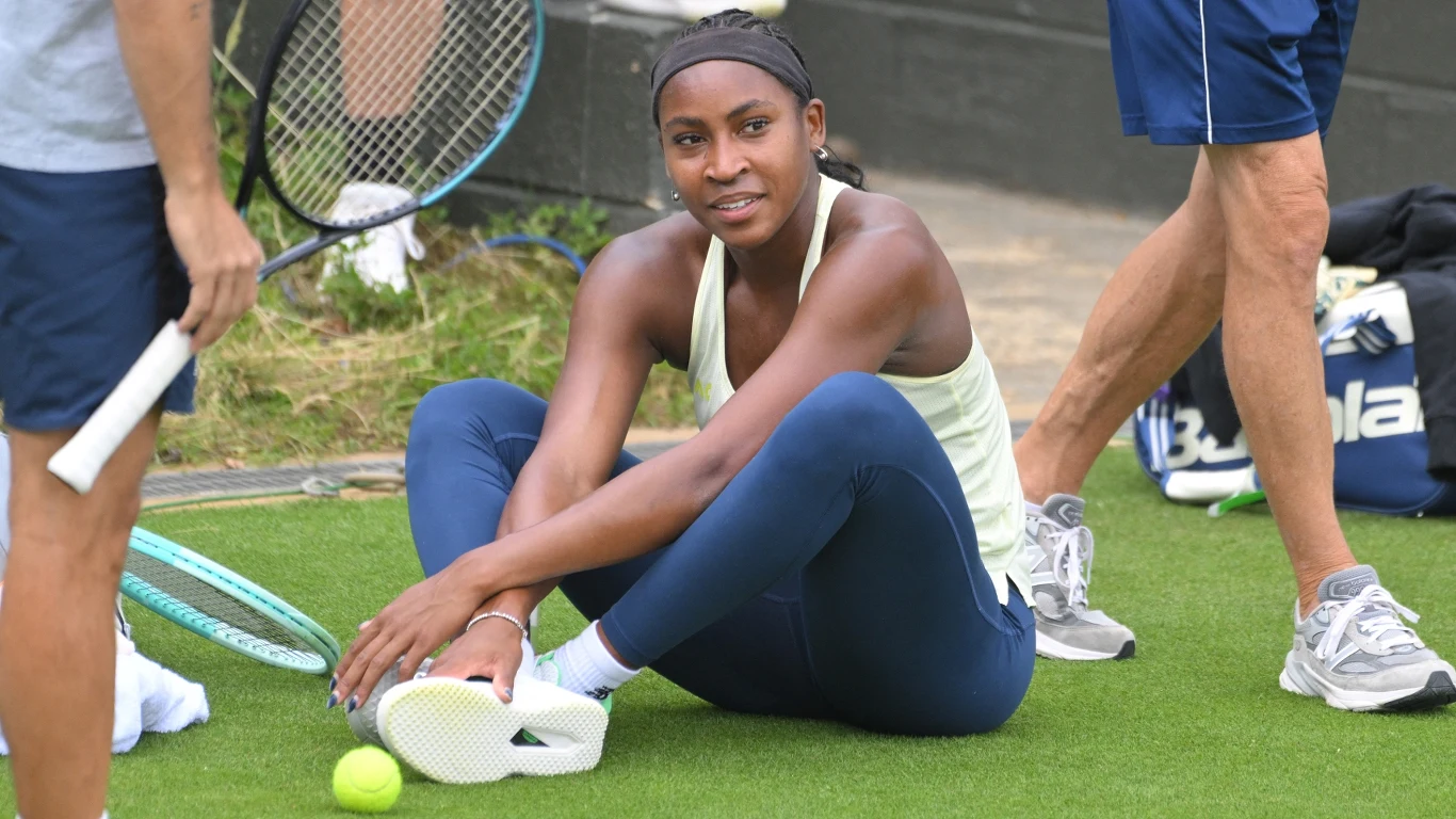 Młoda tenisistka siedzi na trawie podczas przerwy w treningu, otoczona przez członków swojego sztabu oraz rakietę tenisową i piłki. Ubrana w sportowy strój, wygląda na zrelaksowaną i skupioną.