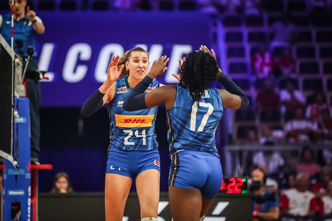 Dwie siatkarki ubrane w niebieskie stroje sportowe świętują zdobycie punktu na boisku, jedna z nich ma wyciągnięte ręce w geście radości; w tle widoczny fragment tablicy wyników oraz trybuny z publicznością.