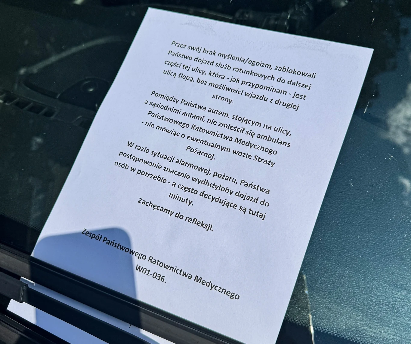 Kartka papieru umieszczona za wycieraczką samochodu z apelem od zespołu ratownictwa medycznego o nieblokowanie dojazdu dla służb ratunkowych, tekst ostrzega przed poważnymi konsekwencjami takiego zachowania.
