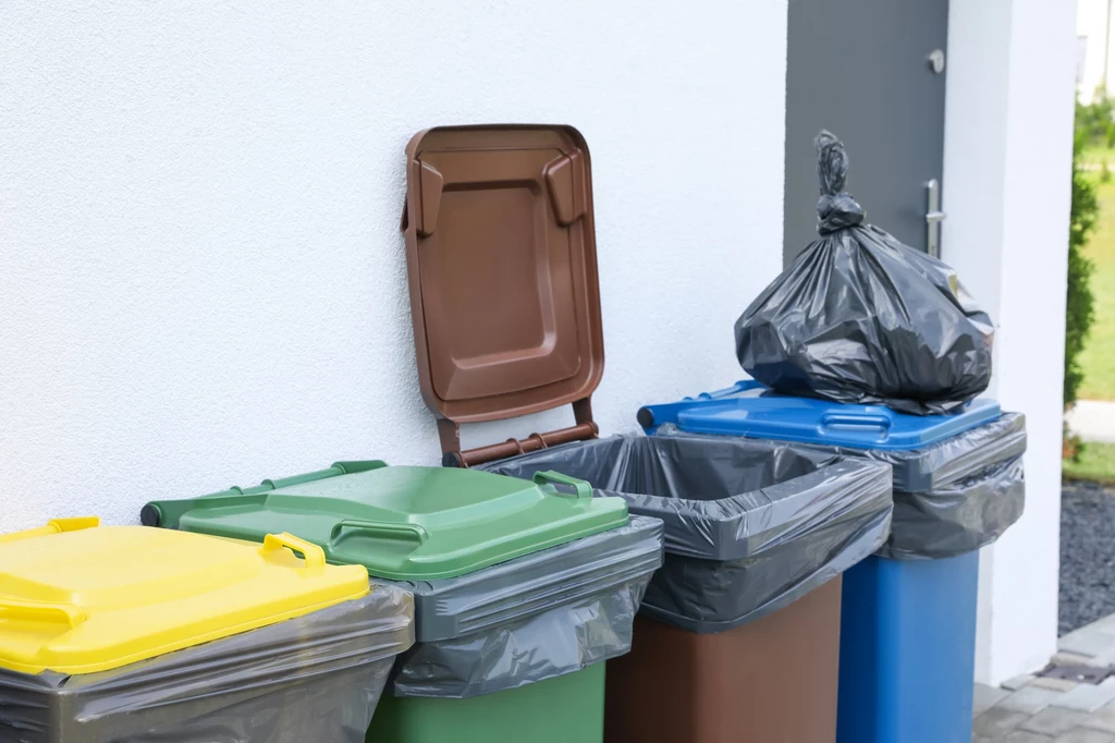 Cztery pojemniki na odpady w różnych kolorach, ustawione przy białej ścianie budynku, każdy przystosowany do segregacji śmieci; na jednym z nich leży czarny worek na śmieci.