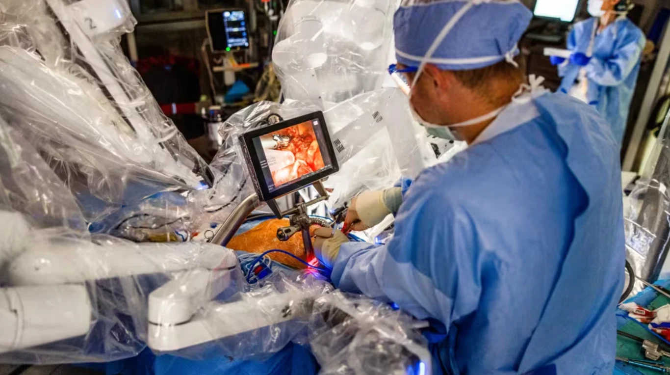 Lekarz w sterylnej odzieży operacyjnej obsługujący robota medycznego, na monitorze wyświetlany jest obraz z wnętrza ciała pacjenta, wokół widoczne są ramiona robota chirurgicznego i sprzęt medyczny.