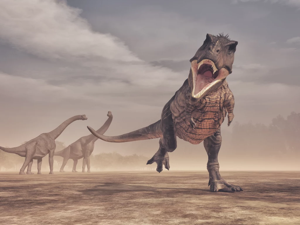 Tyranozaur był silnym mięsożercą, zdolnym do upolowania każdego dinozaura Duży drapieżny dinozaur biegnący z otwartą paszczą na pierwszy planie, w tle dwa roślinożerne dinozaury poruszające się po pustynnym terenie, pochmurne niebo potęguje dramatyczny nastrój sceny.