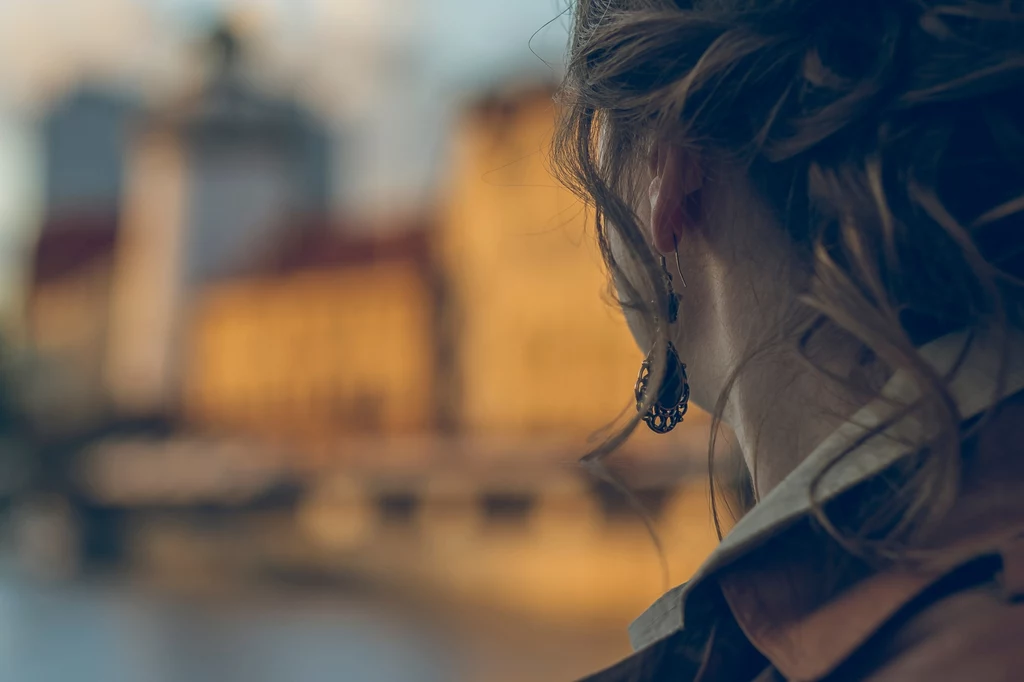 Profil osoby z długimi, kręconymi włosami, patrzącej na rozmazaną panoramę miasta w ciepłym świetle zachodzącego słońca.