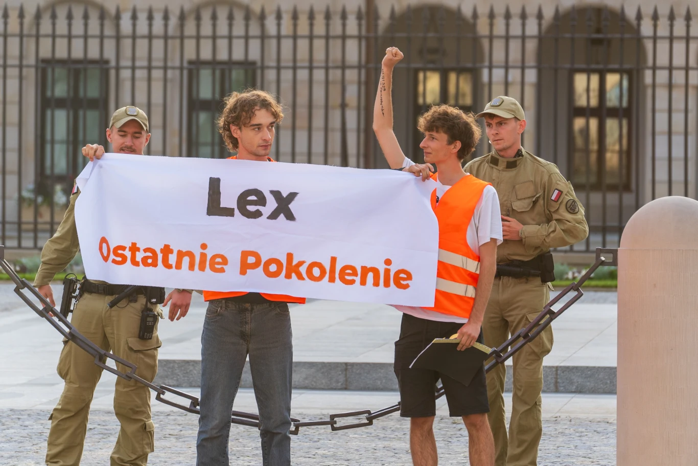 W piątek Ostatnie Pokolenie wdarło się na teren Pałacu Prezydenckiego. Interweniowały służby Dwóch młodych mężczyzn w pomarańczowych kamizelkach trzyma transparent z napisem 'Lex Ostatnie Pokolenie', za nimi stoją dwaj funkcjonariusze w mundurach i czapkach, jeden z protestujących unosi pięść w geście poparcia.