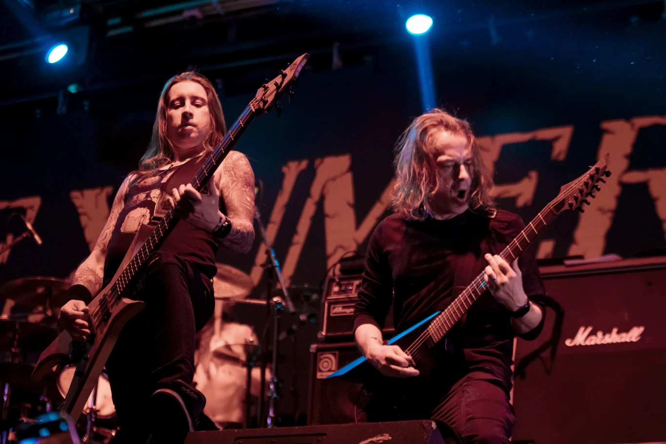 Franz Pries i Bernie Siedler (Exumer) w akcji Dwóch gitarzystów podczas występu na scenie, trzymających gitary elektryczne w dynamicznych pozach, za nimi widoczne oświetlenie sceniczne oraz wzmacniacze, atmosfera koncertu rockowego lub metalowego.