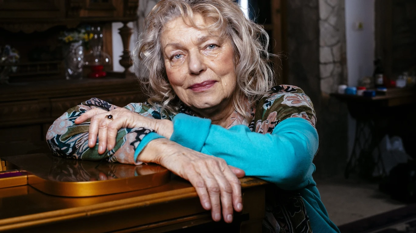 Katarzyna Gaertner (2016) Starsza kobieta o siwych włosach opiera ręce na brązowym fortepianie, patrząc spokojnie przed siebie, otoczona domowym wnętrzem z drewnianymi meblami i nastrojowym światłem.