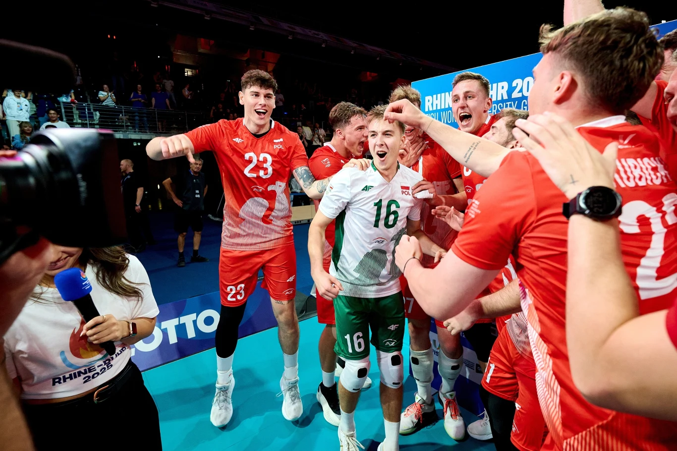 Polscy siatkarze świętujący zwycięstwo w Uniwersjadzie Grupa siatkarzy świętuje na boisku po wygranym meczu, zawodnicy ubrani są w czerwone i białe stroje, wokół panuje radosna atmosfera, a jedna z osób trzyma mikrofon relacjonując wydarzenia.