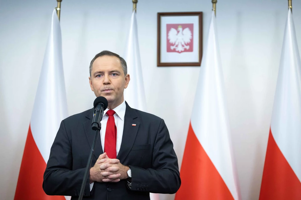 Mężczyzna w garniturze i czerwonym krawacie przemawia do mikrofonu na tle flag Polski oraz godła państwowego zawieszonego na ścianie.