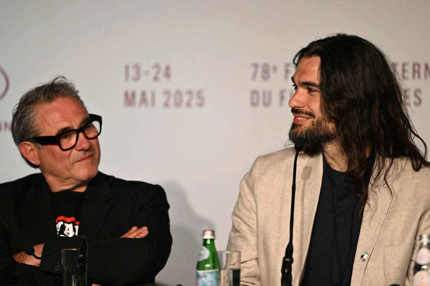 Aktor Sergi Lopez, grający główną rolę i Oliver Laxe - reżyser filmu "Sirat" Dwóch mężczyzn siedzi obok siebie podczas konferencji prasowej, jeden z nich ubrany w czarny garnitur i okulary, drugi z długimi włosami i zarostem w jasnej marynarce; za nimi na ścianie napisy dotyczące daty i miejsca wydarzenia, na stole widoczne mik...