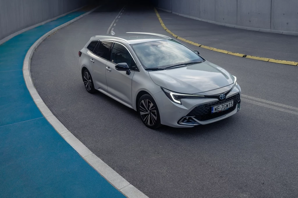 Srebrny samochód osobowy Toyota Corolla kombi poruszający się po wyasfaltowanej drodze otoczonej betonowymi ścianami i niebieskim chodnikiem, widoczny zakręt oraz tunel w tle.