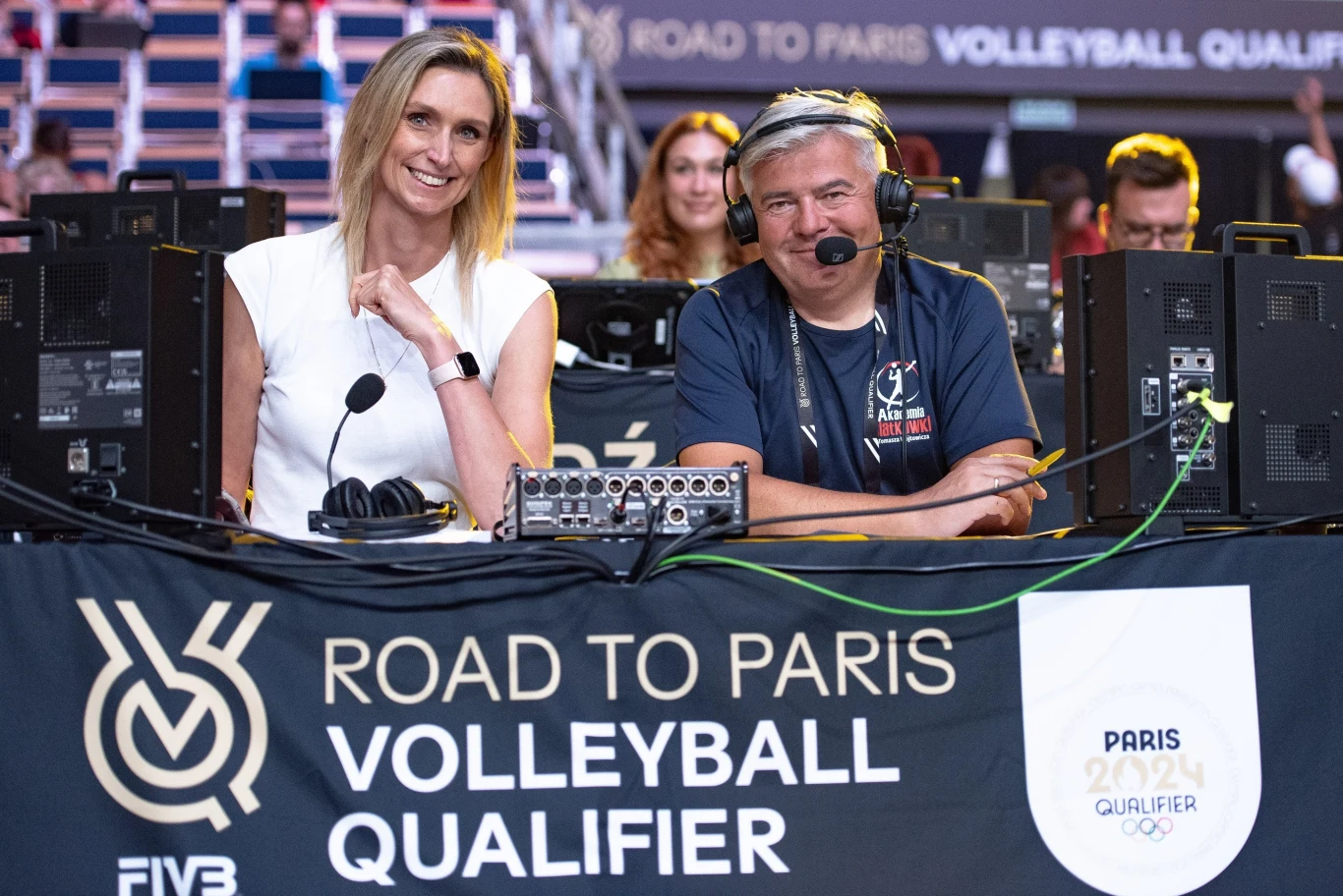 Dwójka komentatorów siedzi za stołem na tle sprzętu audio oraz baneru informującego o eliminacjach do igrzysk olimpijskich siatkówki Paryż 2024, otoczona stadionową atmosferą.