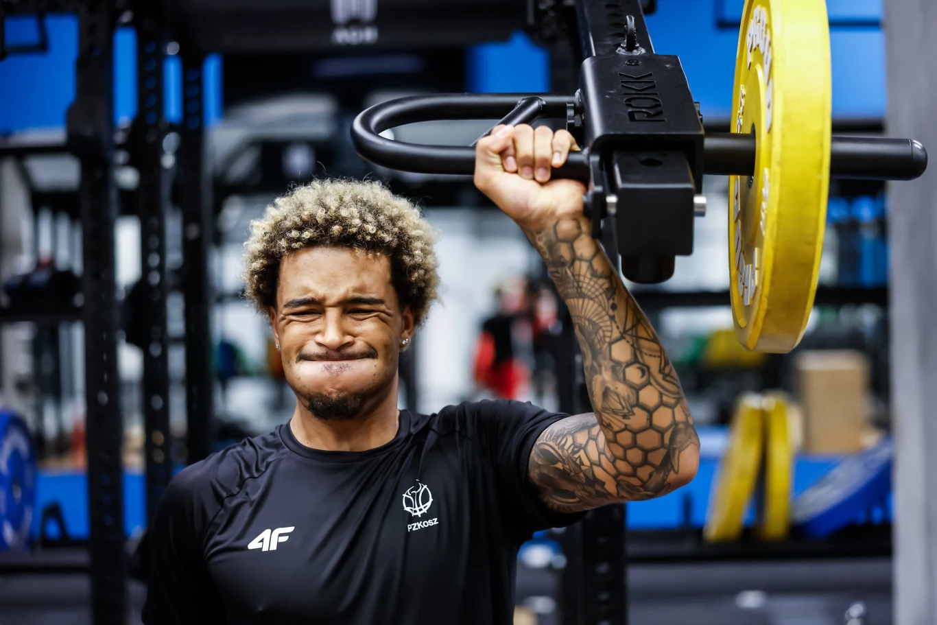 Jeremy Sochan trenuje z kadrą w Krakowie Mężczyzna z blond afro i tatuażem na ręce podnosi ciężar na siłowni, skoncentrowany na wykonywanym ćwiczeniu, ujęcie skupione na jego wysiłku i determinacji.