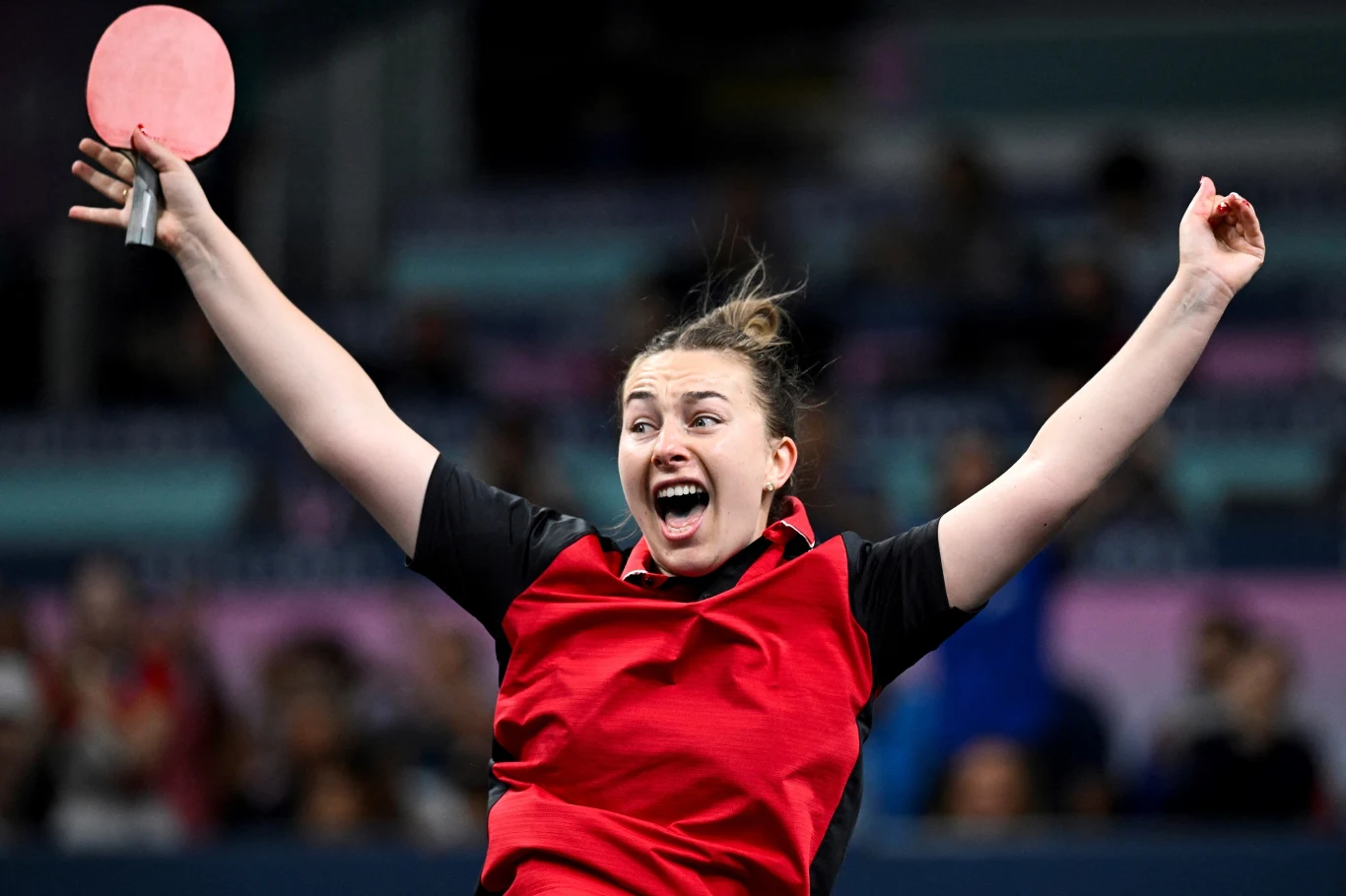 Młoda kobieta w czerwono-czarnym stroju sportowym z uniesionymi rękami oraz szerokim uśmiechem, trzymająca rakietkę do tenisa stołowego, na tle rozmytej publiczności.