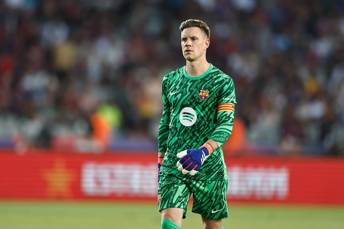 Marc-Andre ter Stegen Bramkarz w zielonym stroju z opaską kapitana na ramieniu stoi na murawie stadionu, w tle niewyraźnie widoczna publiczność oraz reklamy.