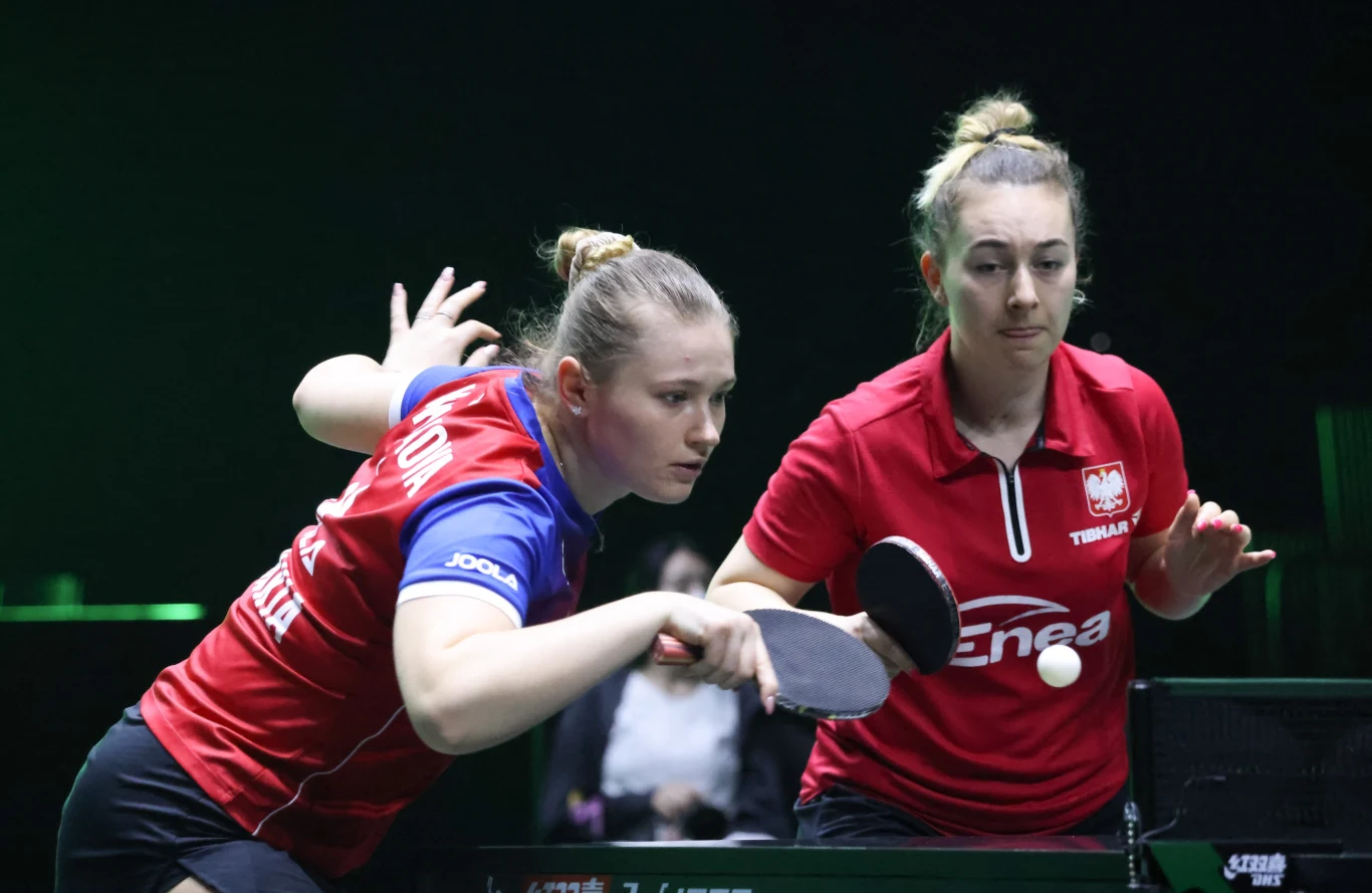 Dwie kobiety w sportowych strojach grają w tenisa stołowego na turnieju, skupione na odbiciu piłeczki i dynamicznej akcji, na tle ciemnego otoczenia.
