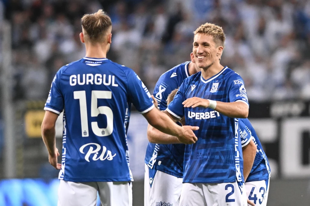 Lech Poznań jest o krok od 3. rundy eliminacji Ligi Mistrzów Dwóch piłkarzy w niebieskich koszulkach Lecha Poznań świętuje na boisku, jeden z nich z uśmiechem gratuluje koledze, w tle widać kibiców na trybunach.