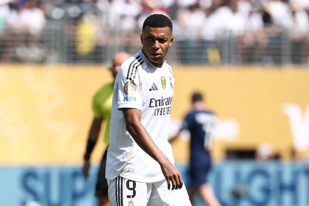 Kylian Mbappe Piłkarz w białym stroju drużyny Realu Madryt podczas meczu piłki nożnej, na tle rozmytego stadionu z kibicami i sędzią w tle.