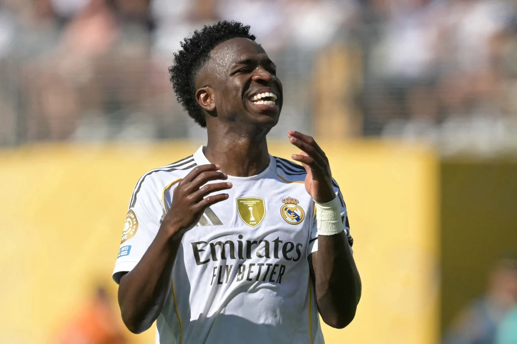 Vinicius Junior Zawodnik w białym stroju klubu Real Madryt cieszy się podczas meczu piłki nożnej, wyrażając radość z gry na boisku, w tle nieostre postacie kibiców i żółta ściana stadionu
