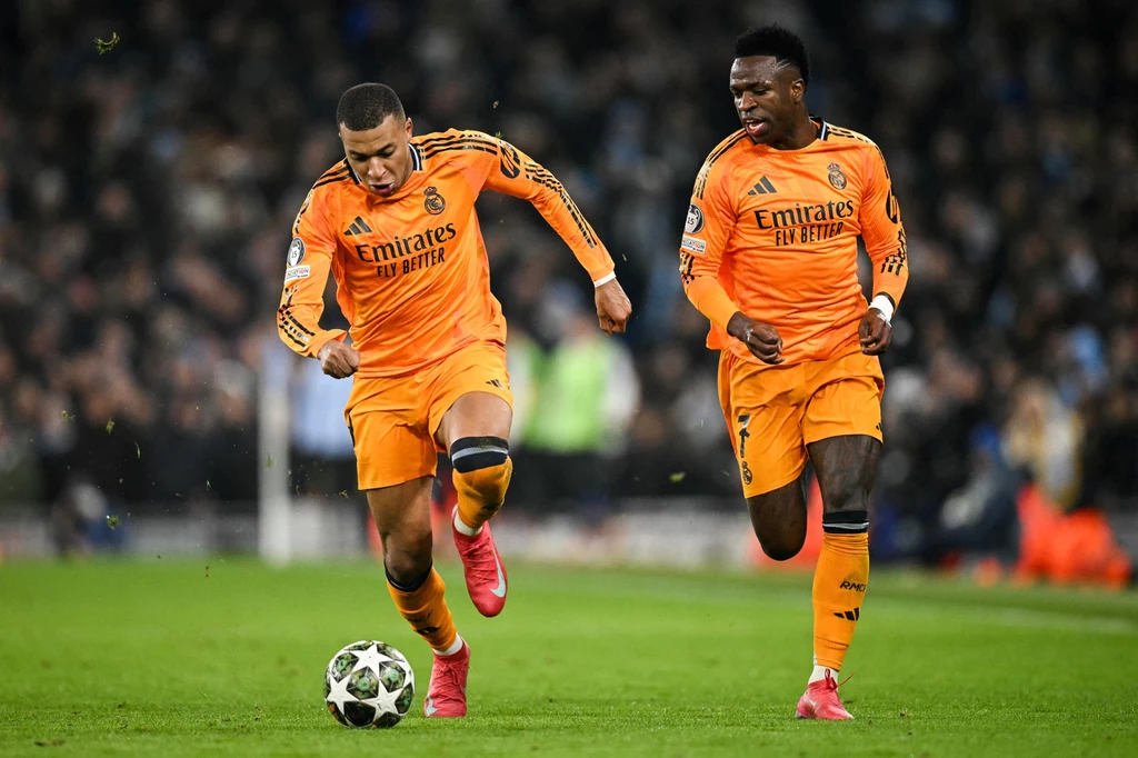 Kylian Mbappe i Vinicius Jr. Dwóch piłkarzy w pomarańczowo-żółtych strojach drużyny Real Madryt biegnie po murawie, jeden z nich prowadzi piłkę podczas meczu, widoczna publiczność w tle.