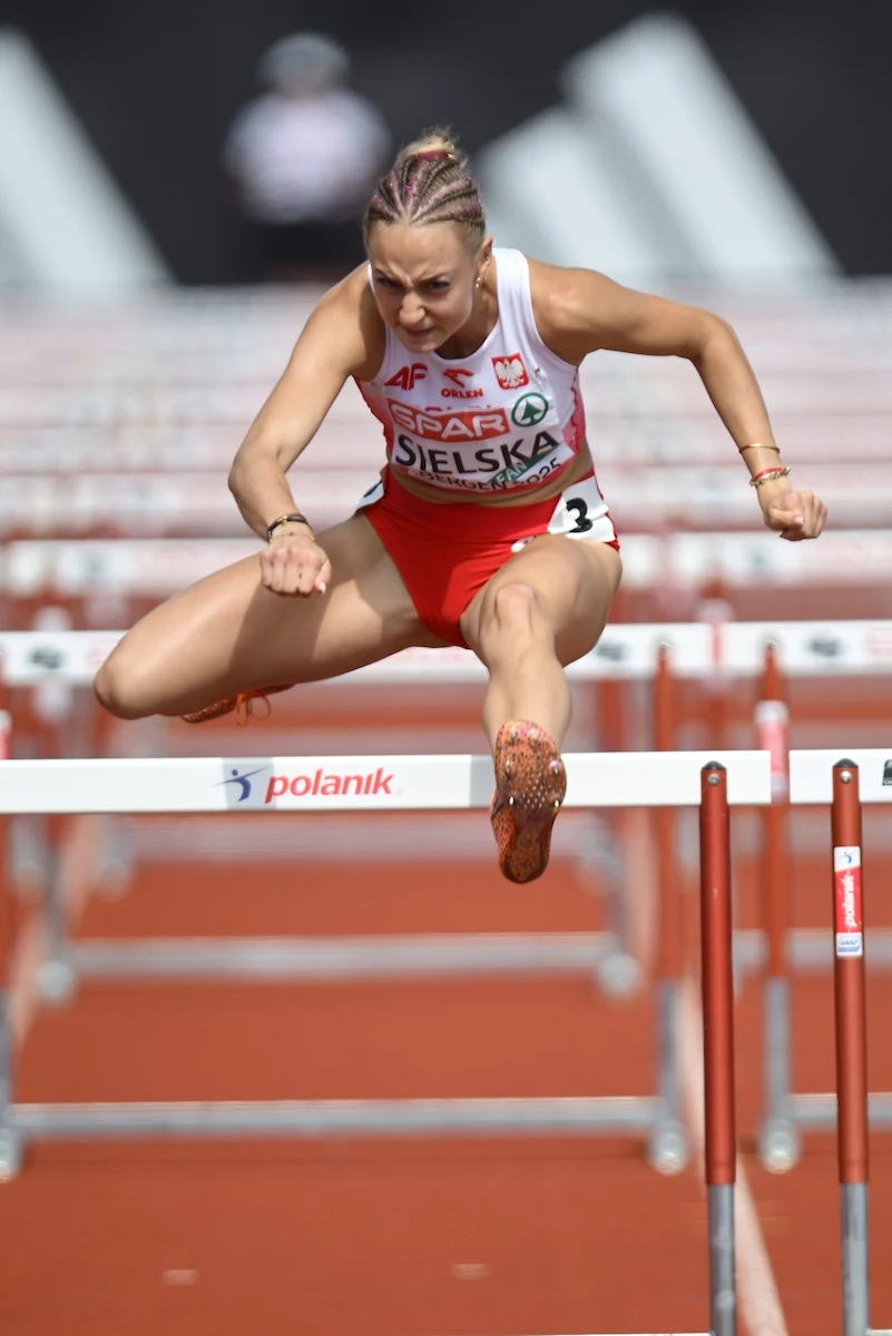 Alicja Sielska Lekkoatletka w biało-czerwonym stroju sportowym przeskakuje przez płotek na bieżni podczas zawodów, skupiona i w dynamicznej pozie, z wyraźnym wysiłkiem na twarzy.