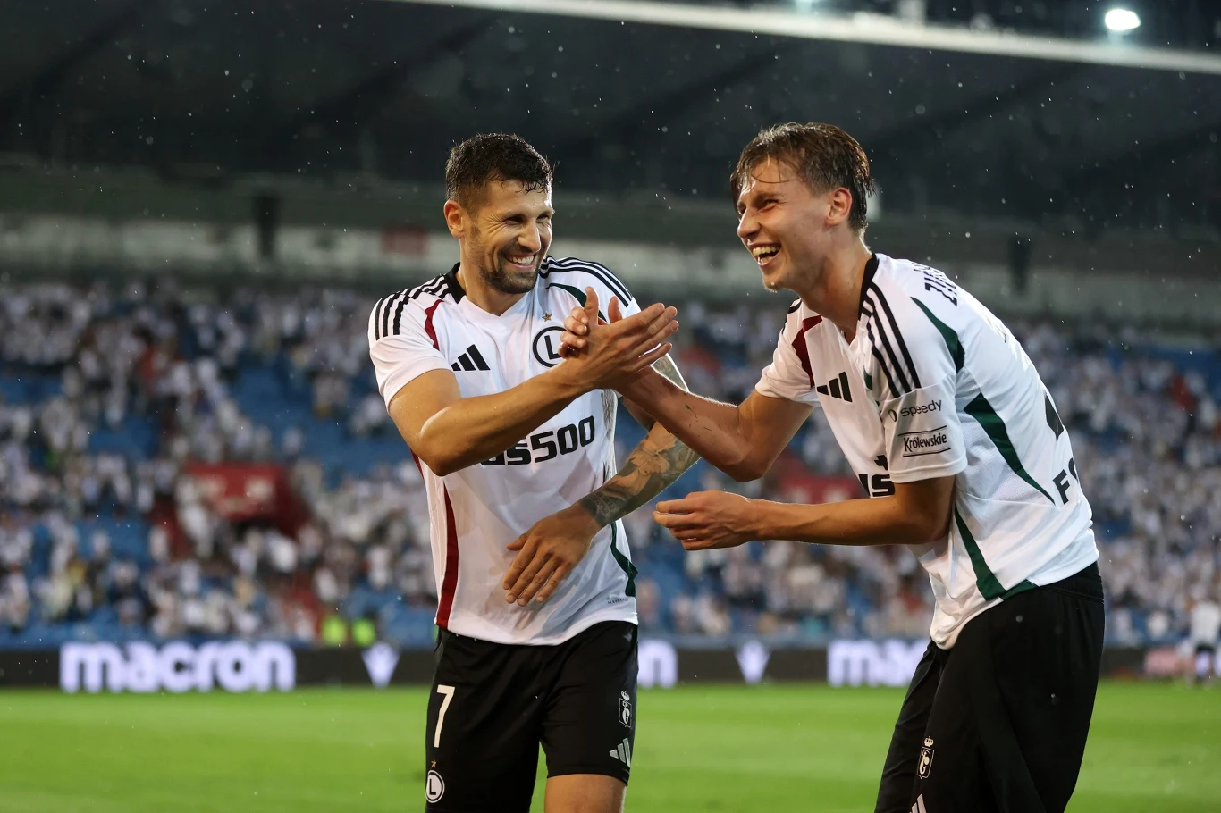 Paweł Wszołek (z lewej) i Jan Ziółkowski na obiekcie Banika Ostrawa Dwóch piłkarzy w białych koszulkach podczas celebracji na stadionie, uśmiechnięci i bijący się dłońmi na tle rozmytej publiczności.