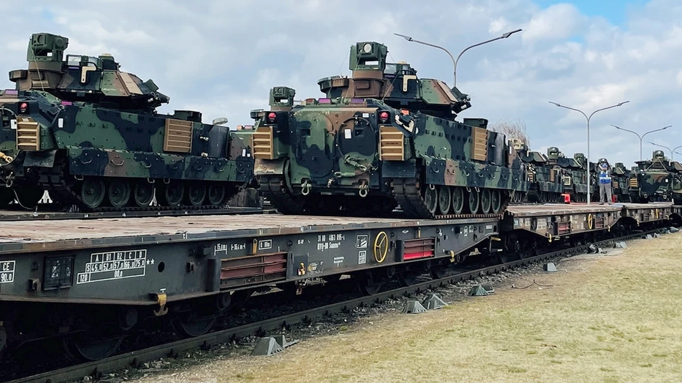 Koszmar Rosjan, wozy bojowe M2 Bradley, już jadą na Ukrainę Transport pojazdów opancerzonych na wagonach kolejowych na stacji, widoczne wozy bojowe z kamuflażem ustawione w rzędzie pod pochmurnym niebem.