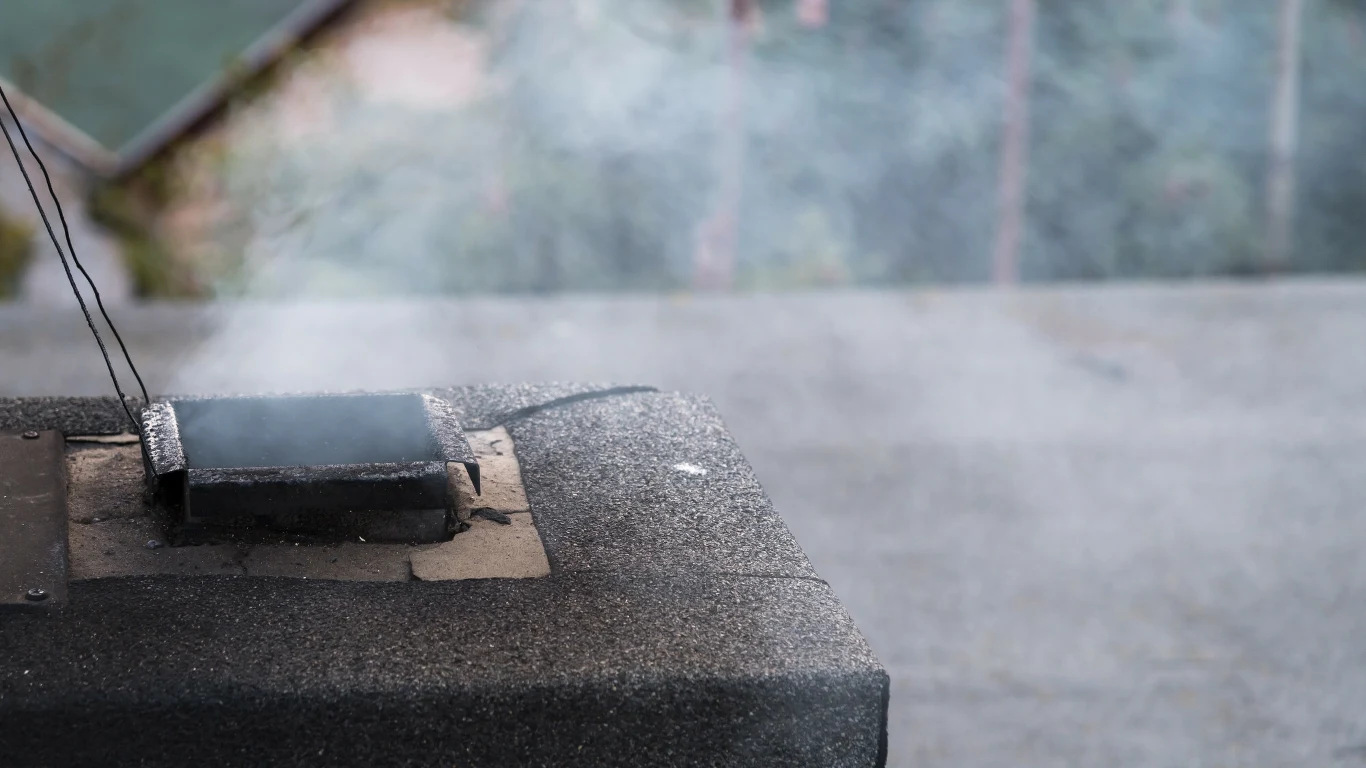 Liczba wniosków składanych w programie "Czyste Powietrze" spadła do zaledwie kilkuset tygodniowo Dym unoszący się z komina wentylacyjnego na dachu, na pierwszym planie widoczne pokrycie dachowe i niewielki otwór z klapą wentylacyjną.