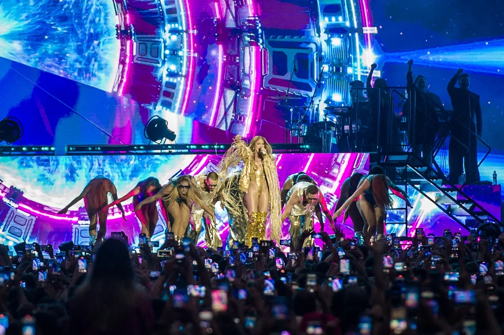 Jennifer Lopez w Warszawie Artystka w złotej kreacji wykonuje utwór na scenie podczas widowiskowego koncertu, otoczona przez grupę tancerzy oraz kolorowe efekty świetlne i wizualizacje, tłum fanów z telefonami rejestruje występ.