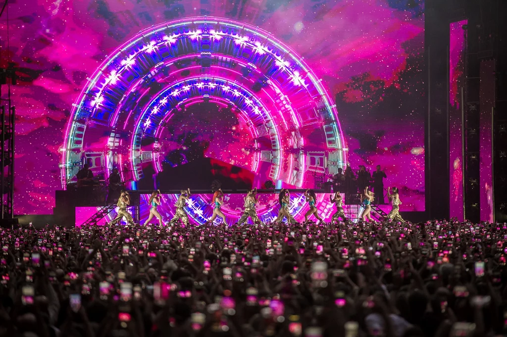 Jennifer Lopez w Warszawie Kolorowa scena koncertowa z efektem świetlnym w kształcie tęczy na tle, na scenie występują tancerki, a tłum ludzi przed sceną nagrywa wydarzenie telefonami.