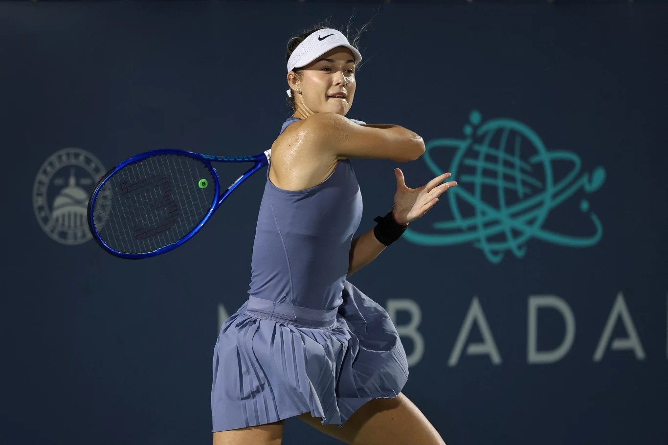 Anna Kalinska Tenisistka wykonująca energiczny zamach rakietą podczas meczu, ubrana w sportową sukienkę, na tle niebieskiej ściany z logo turnieju Mubadala.