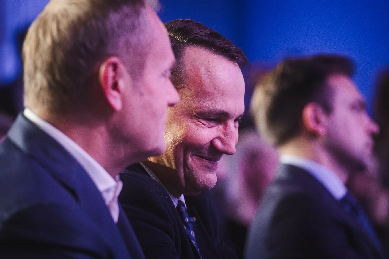 Trzech mężczyzn w garniturach siedzi obok siebie, jeden z nich uśmiecha się szeroko, a reszta zachowuje poważny wyraz twarzy, tło rozmyte w niebieskich tonach.