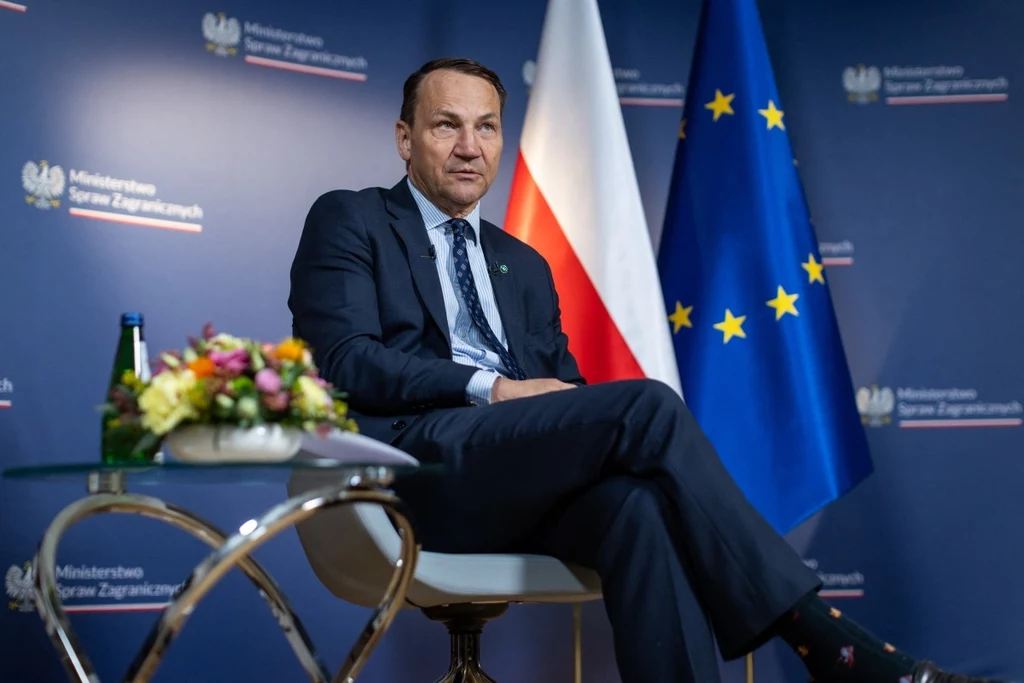 Wicepremier oraz szef MSZ Radosław Sikorski Mężczyzna w garniturze siedzący na krześle w nowoczesnym wnętrzu na tle flag Polski oraz Unii Europejskiej, obok stolik z butelką wody i bukietem kwiatów, ściana z logotypami Ministerstwa Spraw Zagranicznych.