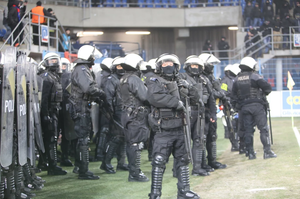 Grupa uzbrojonych i wyposażonych w kaski oraz tarcze policjantów oddziałów prewencji stojących na płycie stadionu w oczekiwaniu na rozwój wydarzeń, w tle trybuny i fragmenty widowni.