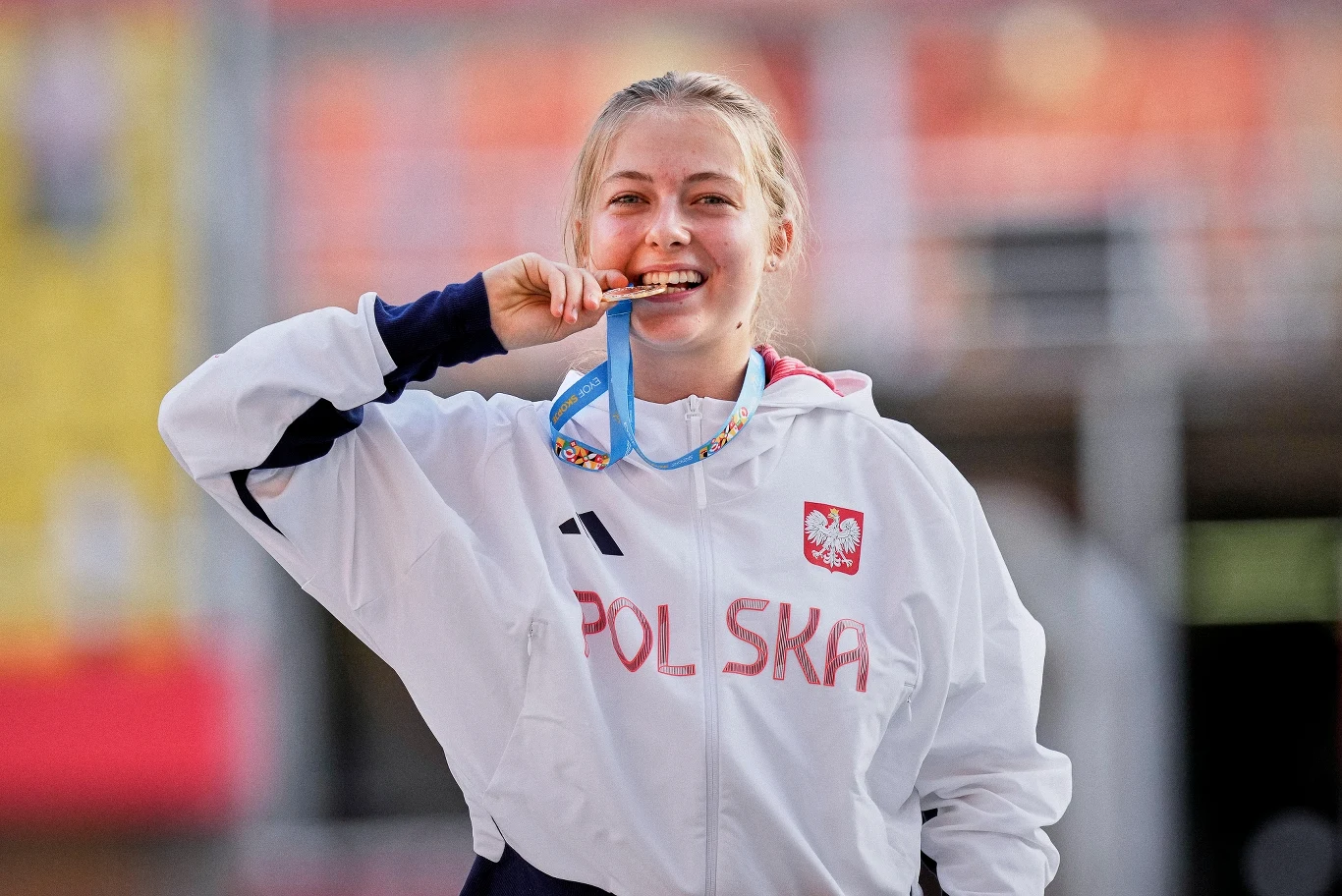 Młoda kobieta w białej sportowej kurtce z napisem 'Polska' oraz godłem, uśmiecha się i gryzie medal zawieszony na niebieskiej wstążce, stojąc na tle stadionu.