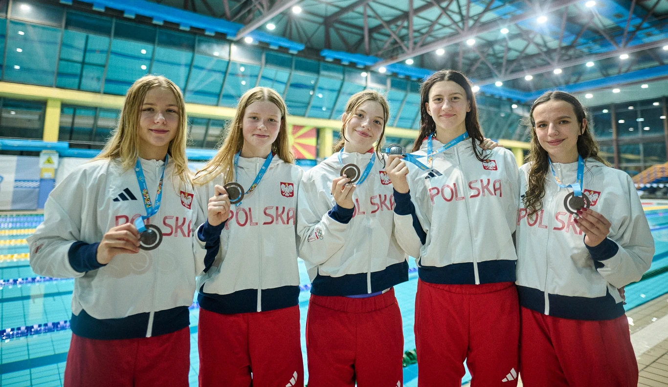 Grupa pięciu młodych zawodniczek w biało-czerwonych strojach sportowych z napisem Polska, stojących na tle krytego basenu, każda z nich trzyma medal na niebieskiej wstążce i uśmiecha się do aparatu.