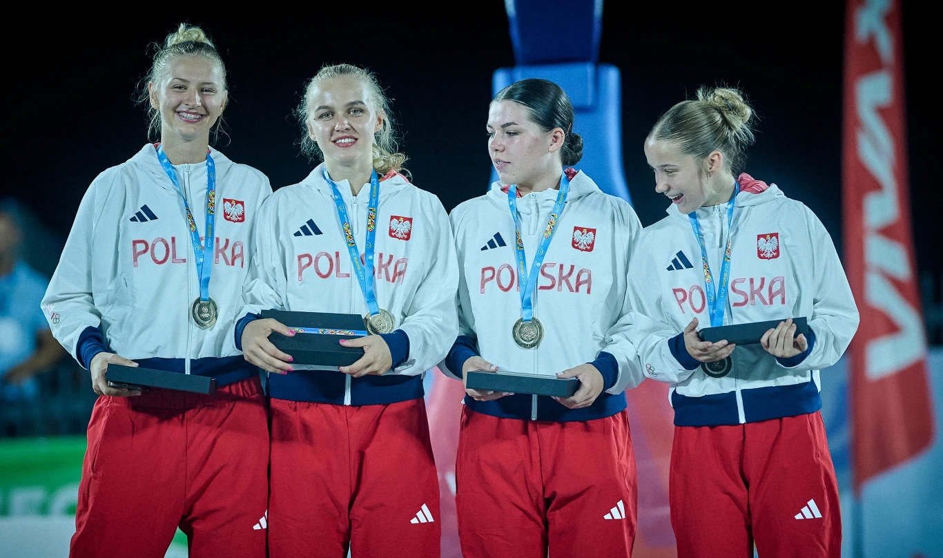Cztery młode kobiety w sportowych biało-czerwonych dresach z napisem POLSKA, każda z medalem na szyi i dekoracyjnym pudełkiem w ręce, stoją obok siebie na tle wieczornego oświetlenia, uśmiechając się i rozmawiając.