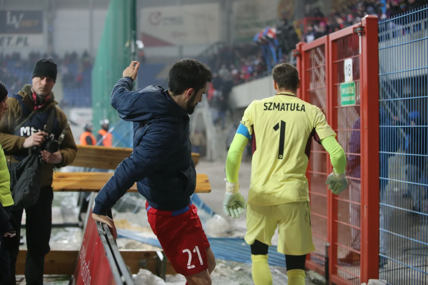 Gerard Badia (po lewej) i Jakub Szmatuła w drodze na rozmowę z kibicami Piasta. Dwóch piłkarzy na tle trybun pełnych kibiców, jeden ubrany w żółty strój bramkarza z napisem 'SZMATUŁA' opuszcza boisko, drugi w czerwonych spodenkach i niebieskiej kurtce wykonuje dynamiczny ruch przy barierek.