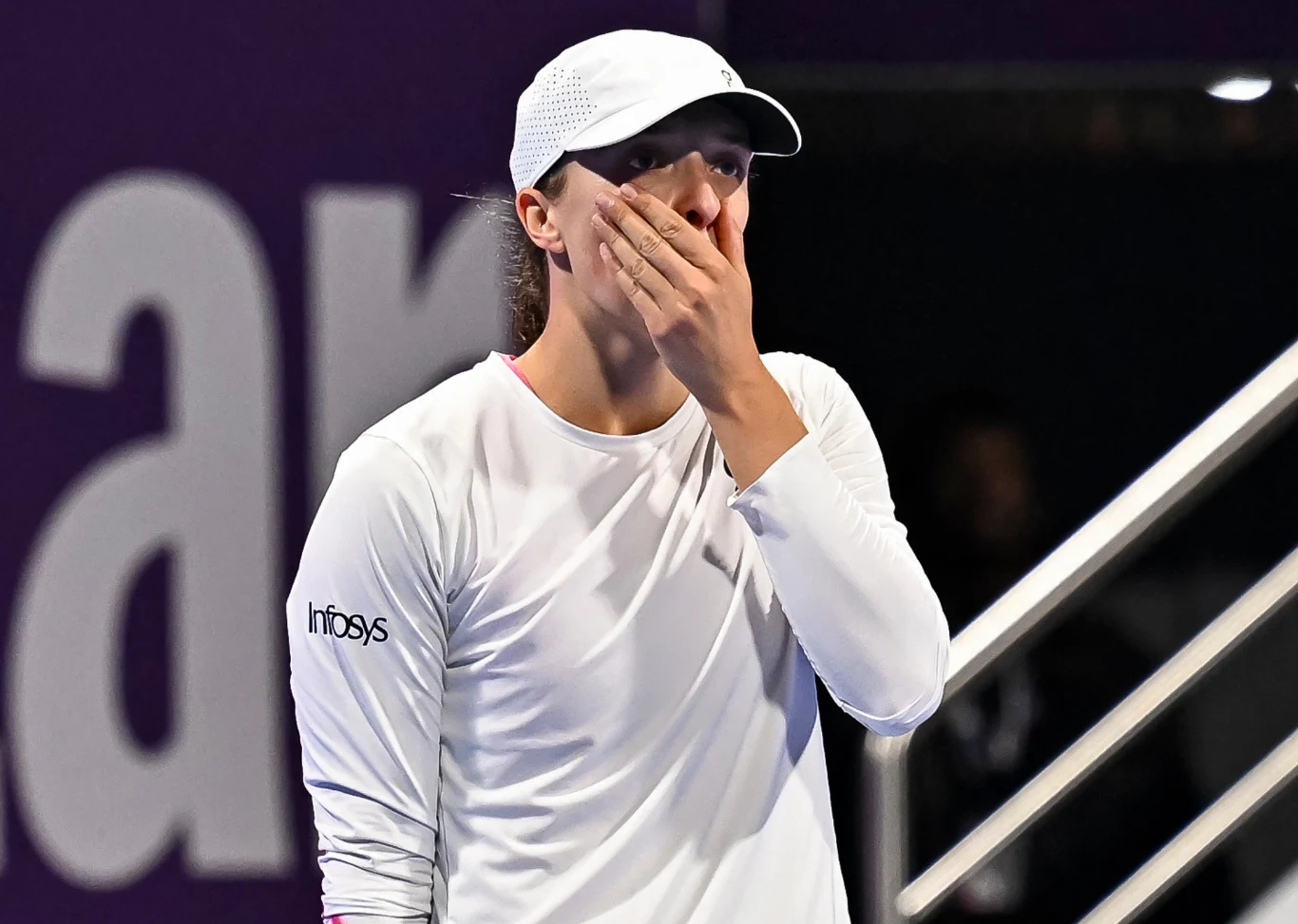 Iga Świątek. Była i - miejmy nadzieję - także przyszła liderka rankingu WTA Sportowiec ubrany na biało, z białą czapką na głowie, zakrywa usta dłonią w geście zaskoczenia lub emocji; w tle widać fragment balustrady i rozmyte napisy.