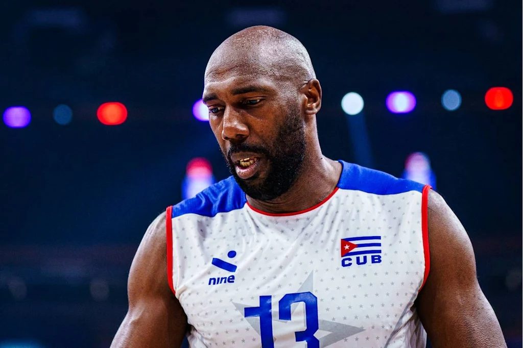 Robertlandy Simon Sportowiec w koszulce drużyny siatkarskiej z numerem 13 i flagą Kuby, skoncentrowany przed meczem lub w trakcie zawodów, na tle rozmytego oświetlenia hali sportowej.